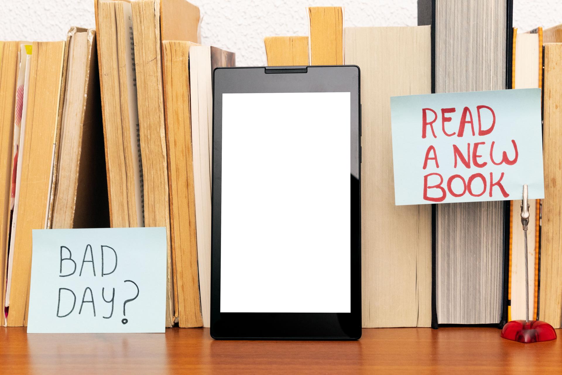 Tablet with blank screen resting on a row of books on a wooden shelf; two notes reading Bad day? and Read a new book stand out. Concept of traditional and electronic reading.